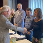 Ein Mann schüttelt einer Frau die Hand. Im Hintergrund beobachten mehrere Personen dies.